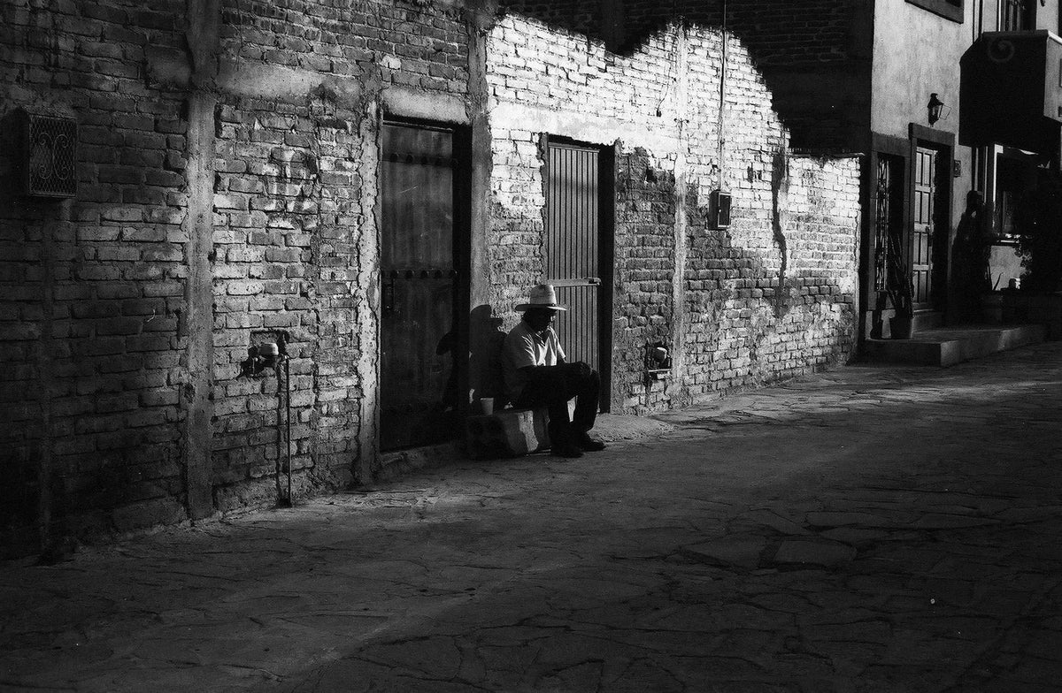 A person sits in shadow beside a sunlit brick wall on a narrow street.