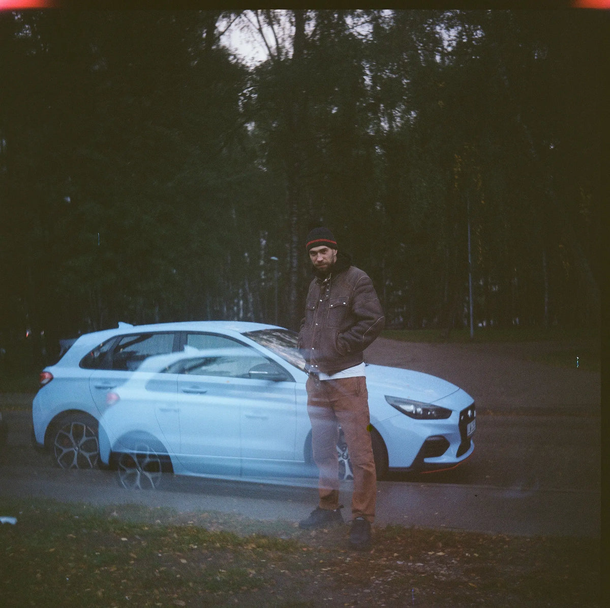 A man standing beside a pale blue car at the edge of trees.
