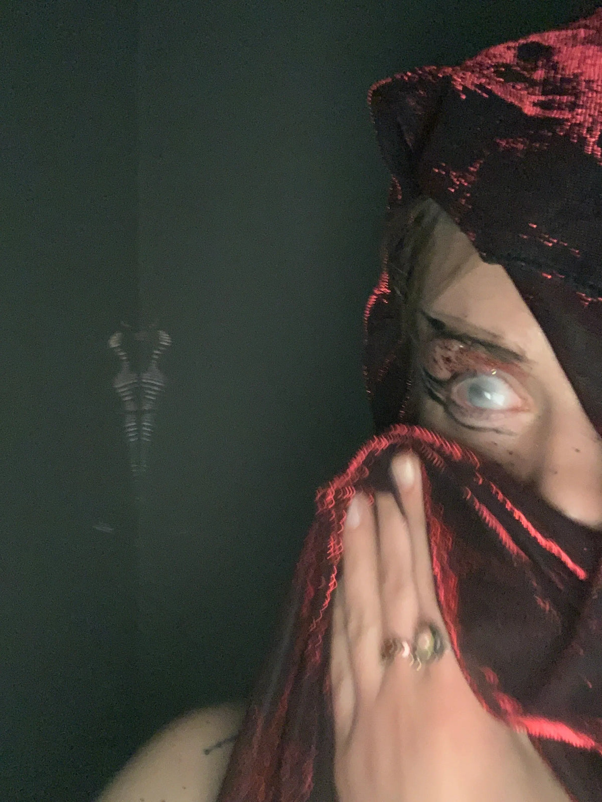 A close self-portrait with one pale eye, a hand and red-edged dark fabric against a dark background.